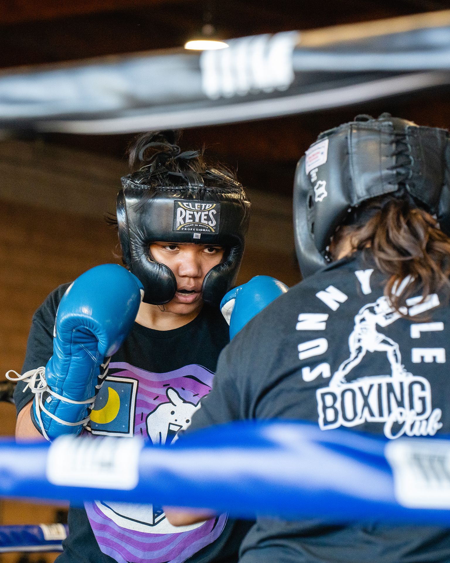 Two boxers sparring in a ring. One with blue gloves and headgear; the other in the background with back turned.