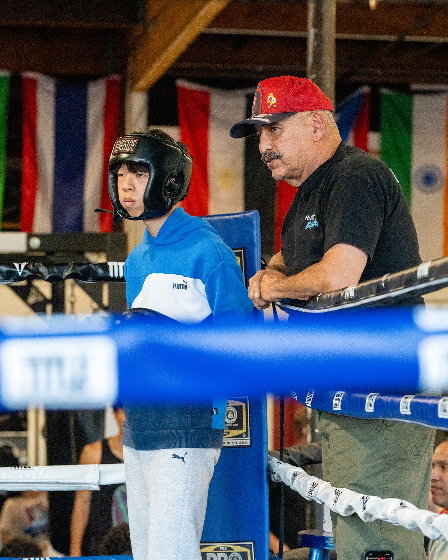 Boxer in helmet, coach in red cap, leaning on ring ropes, flags in background.
