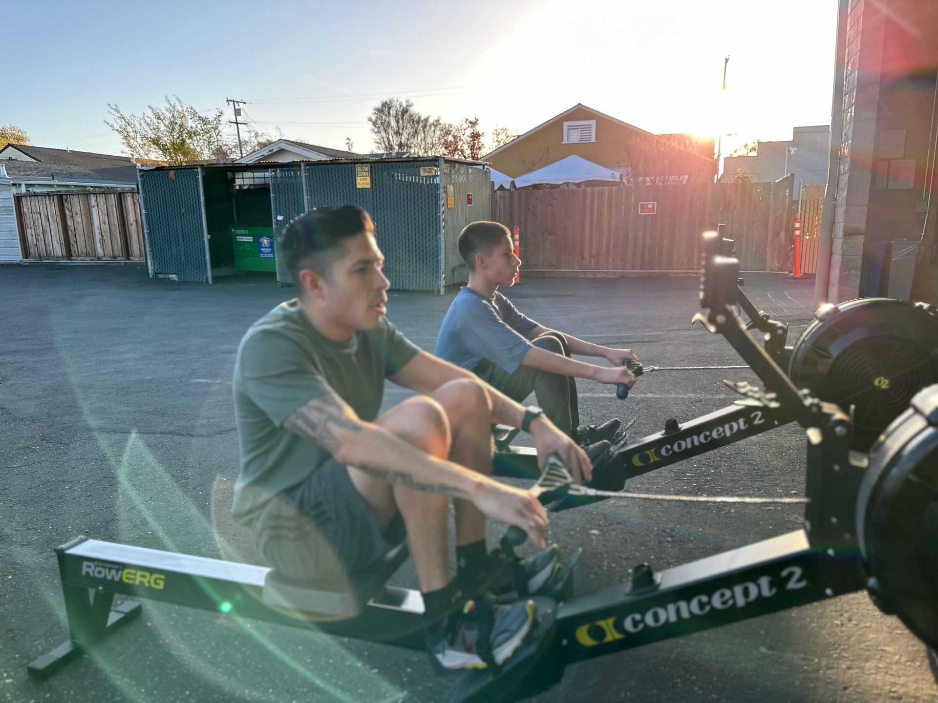 Two men rowing on Concept 2 machines outside, sun setting in background.