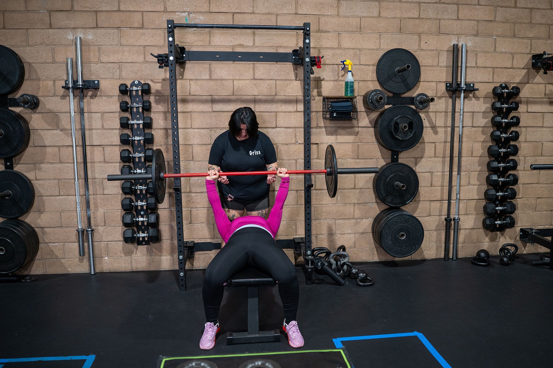 Woman bench presses with spotter in a gym. Both wear black and pink workout clothes. Brick wall, weights.