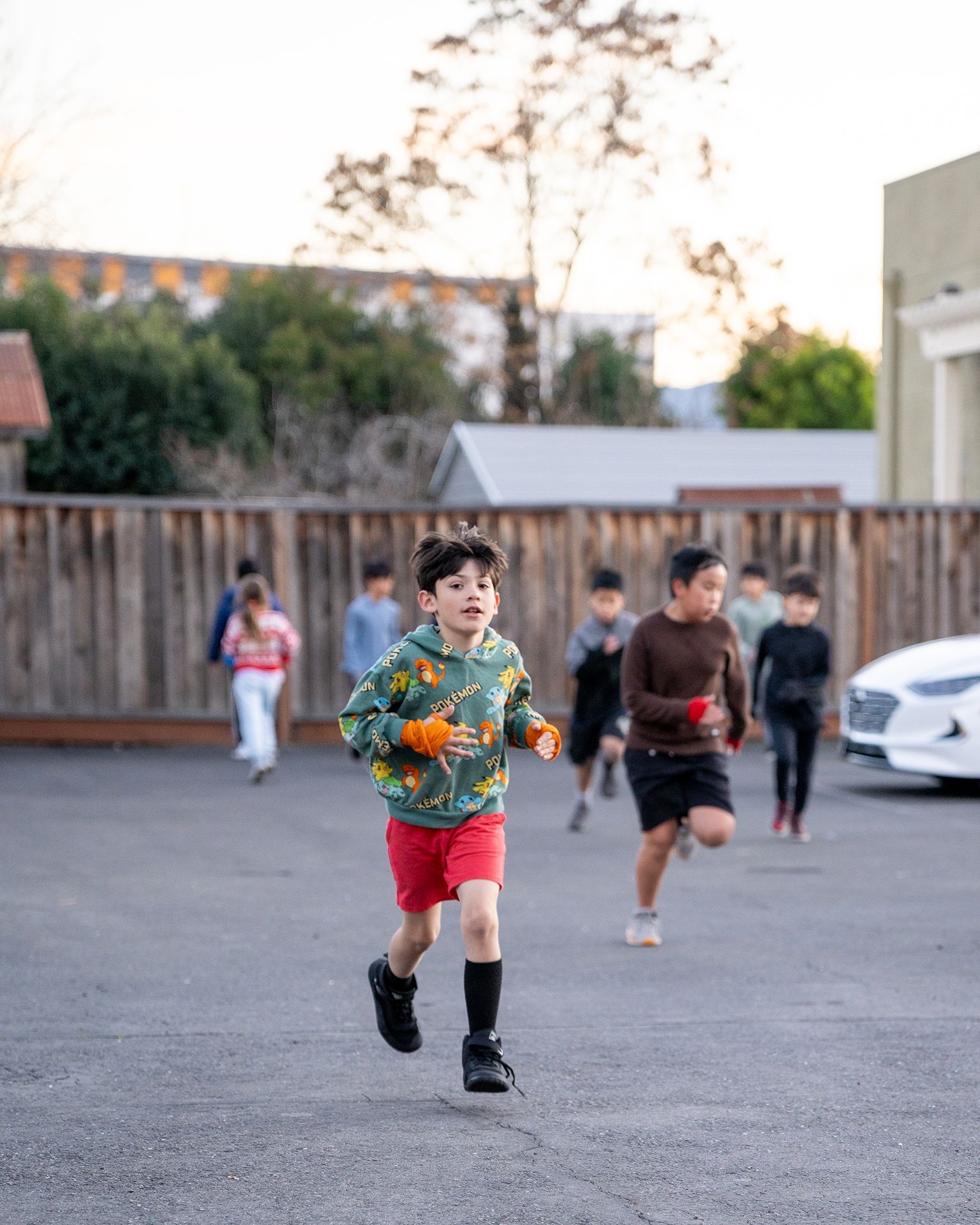 Group of children running on a paved area near a wooden fence and buildings. One child in the foreground is smiling.