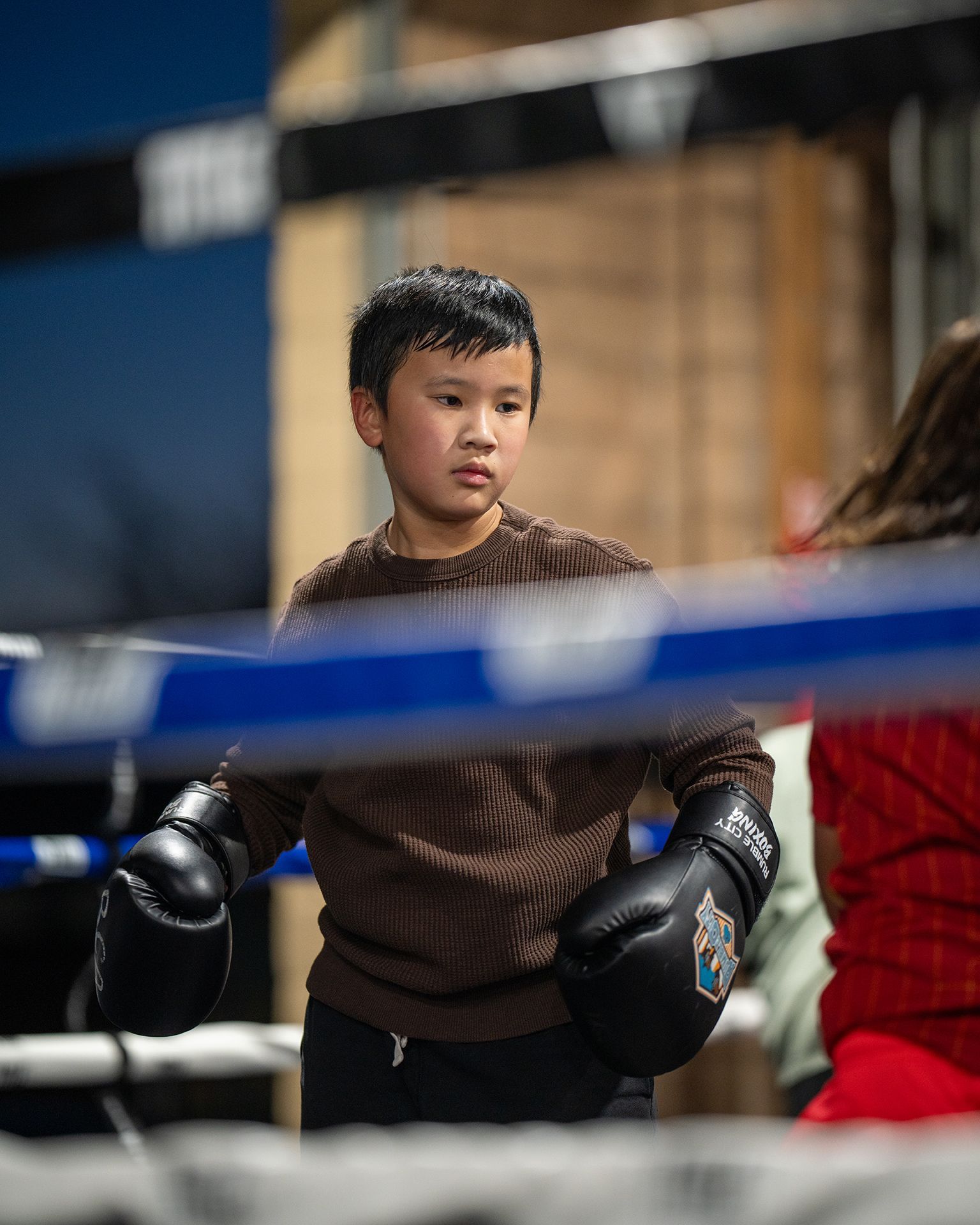 Young boy in boxing gloves stands inside a boxing ring, looking focused.
