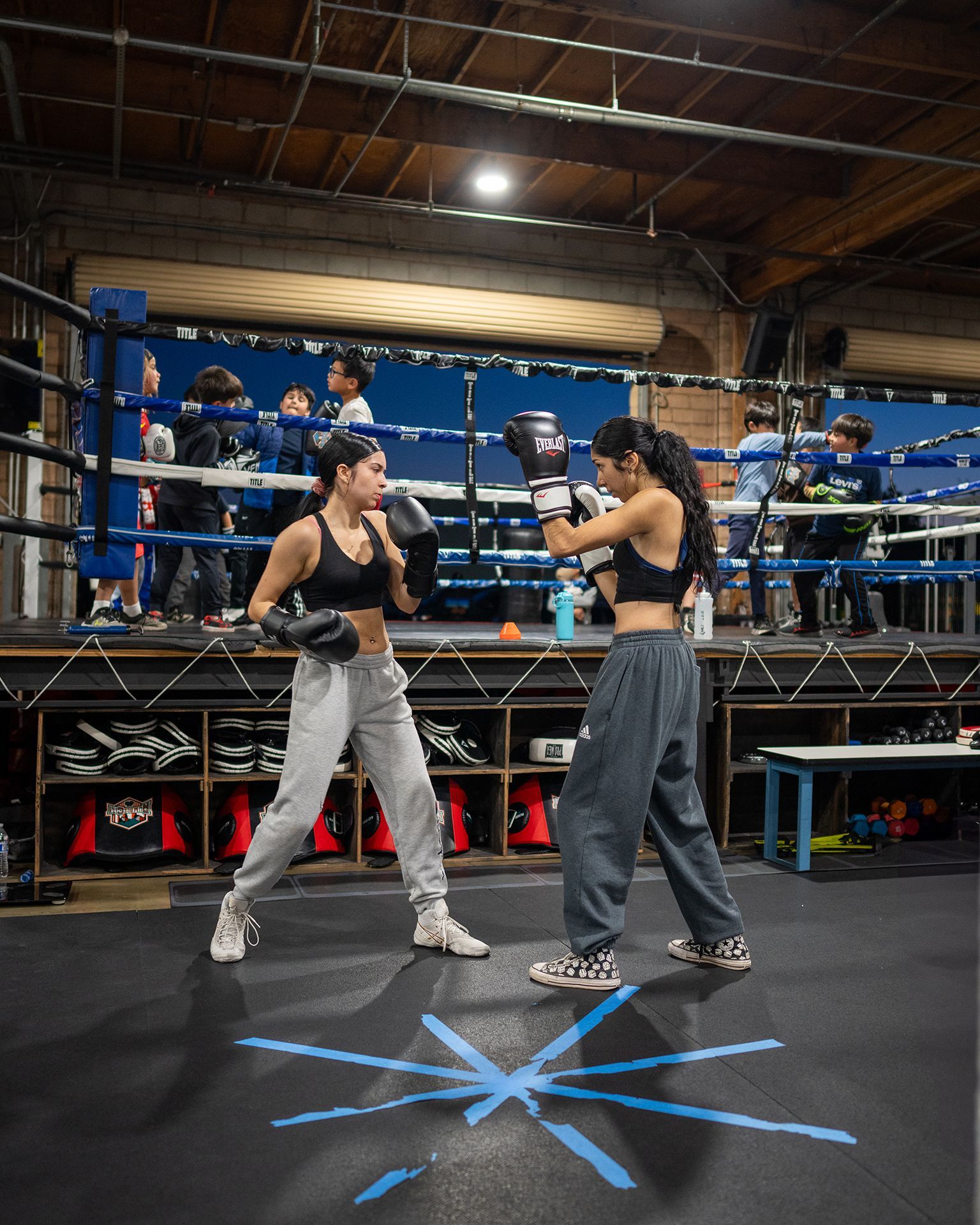 Two women boxing in a ring, wearing sportswear and gloves. People watch in the background of the gym.