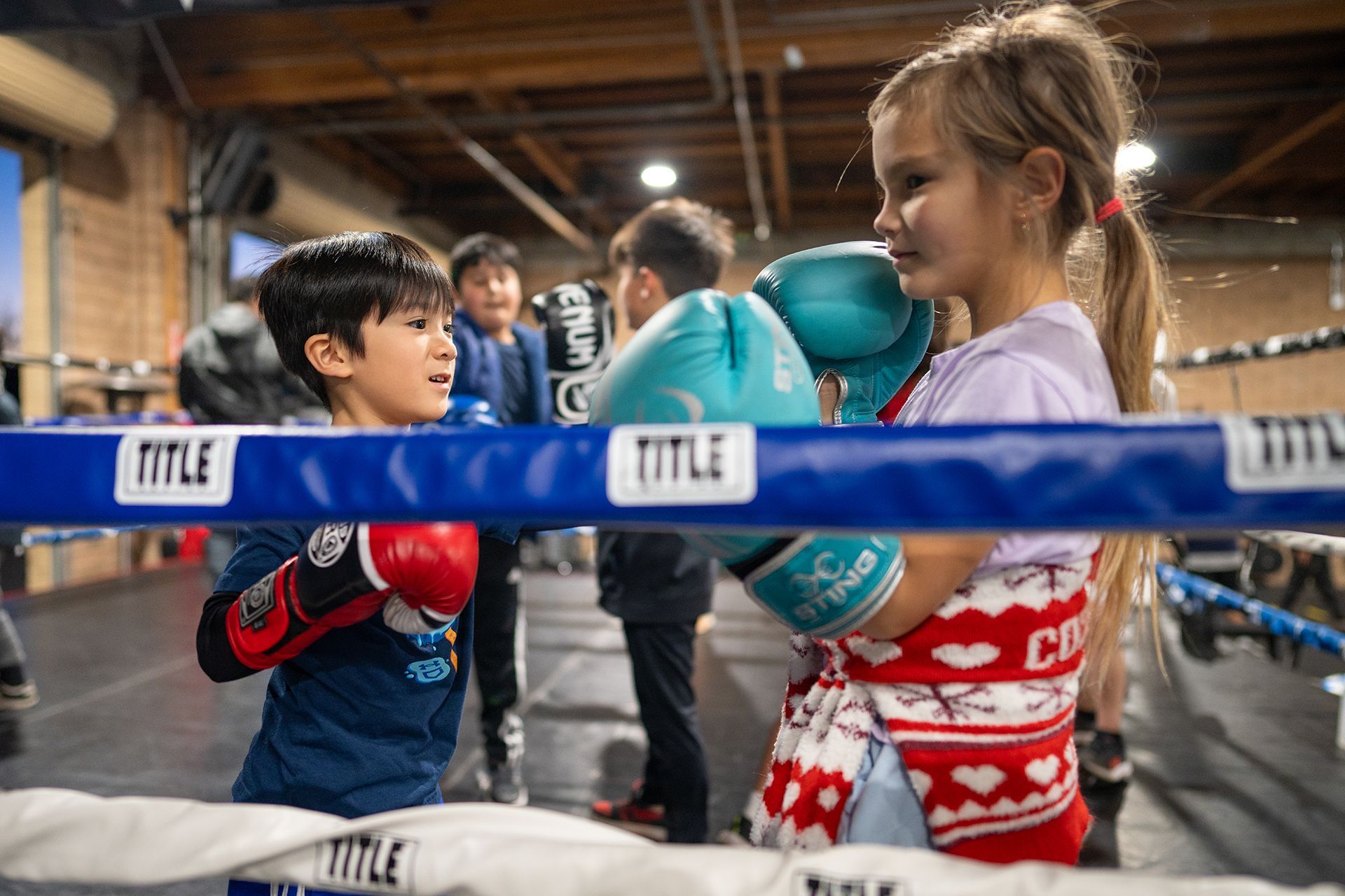Two children in boxing gloves sparring inside a boxing ring; other kids and adults in the background.