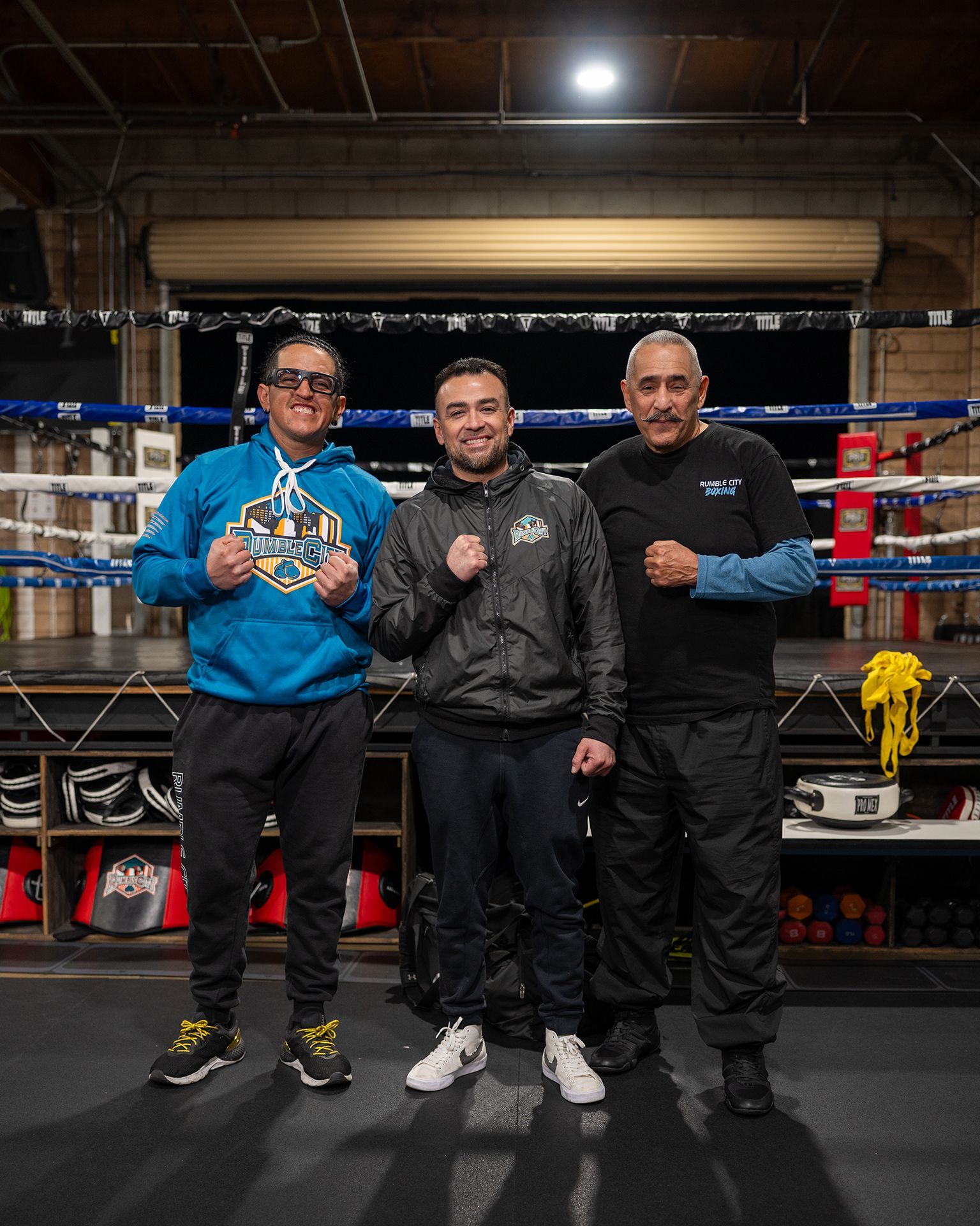 Three men pose in a boxing ring, fists up. One wears blue, another black jacket, the last a black shirt.