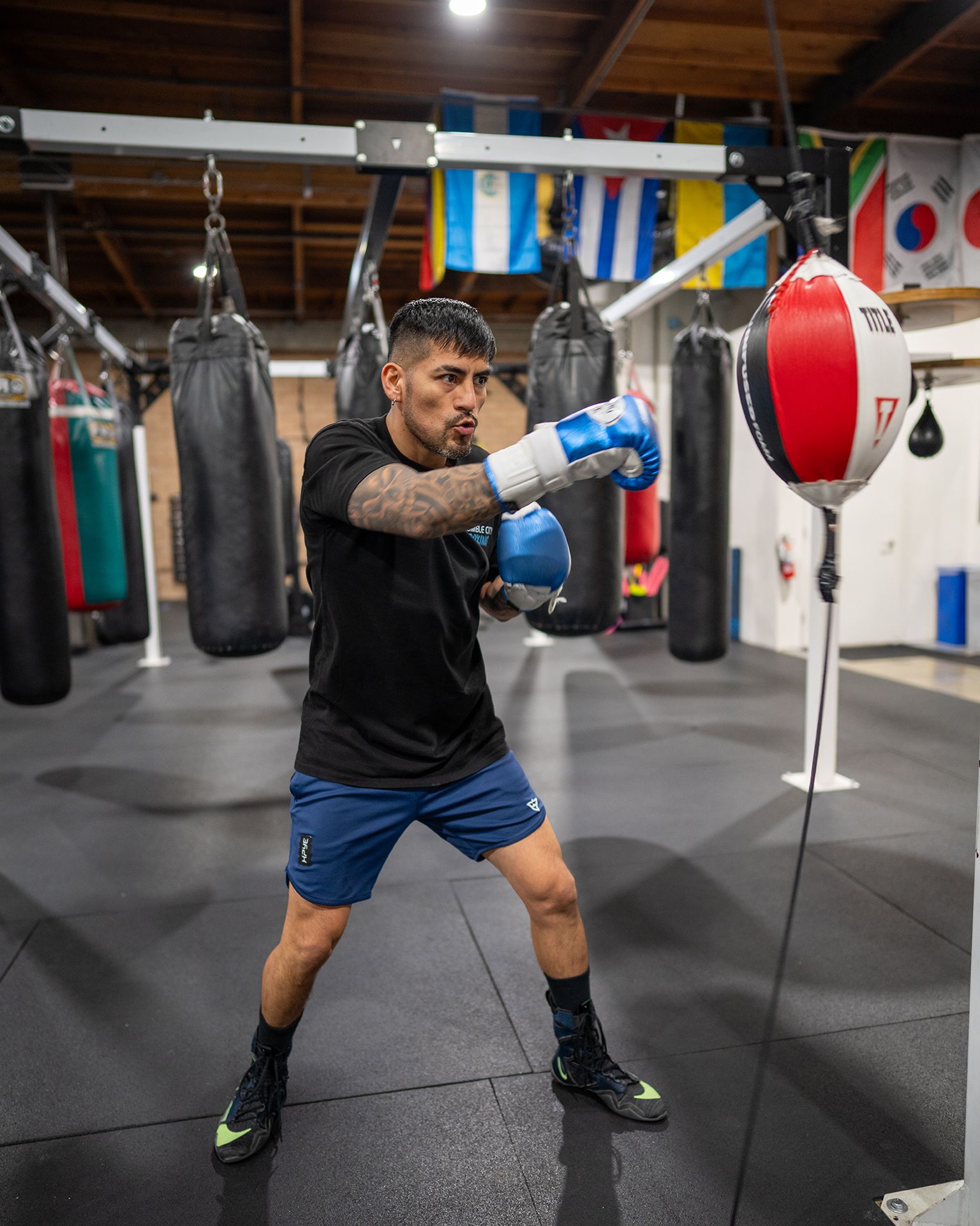 Boxer in gym throwing a punch at a speed bag; wearing gloves, shorts, tattoos, near flags.