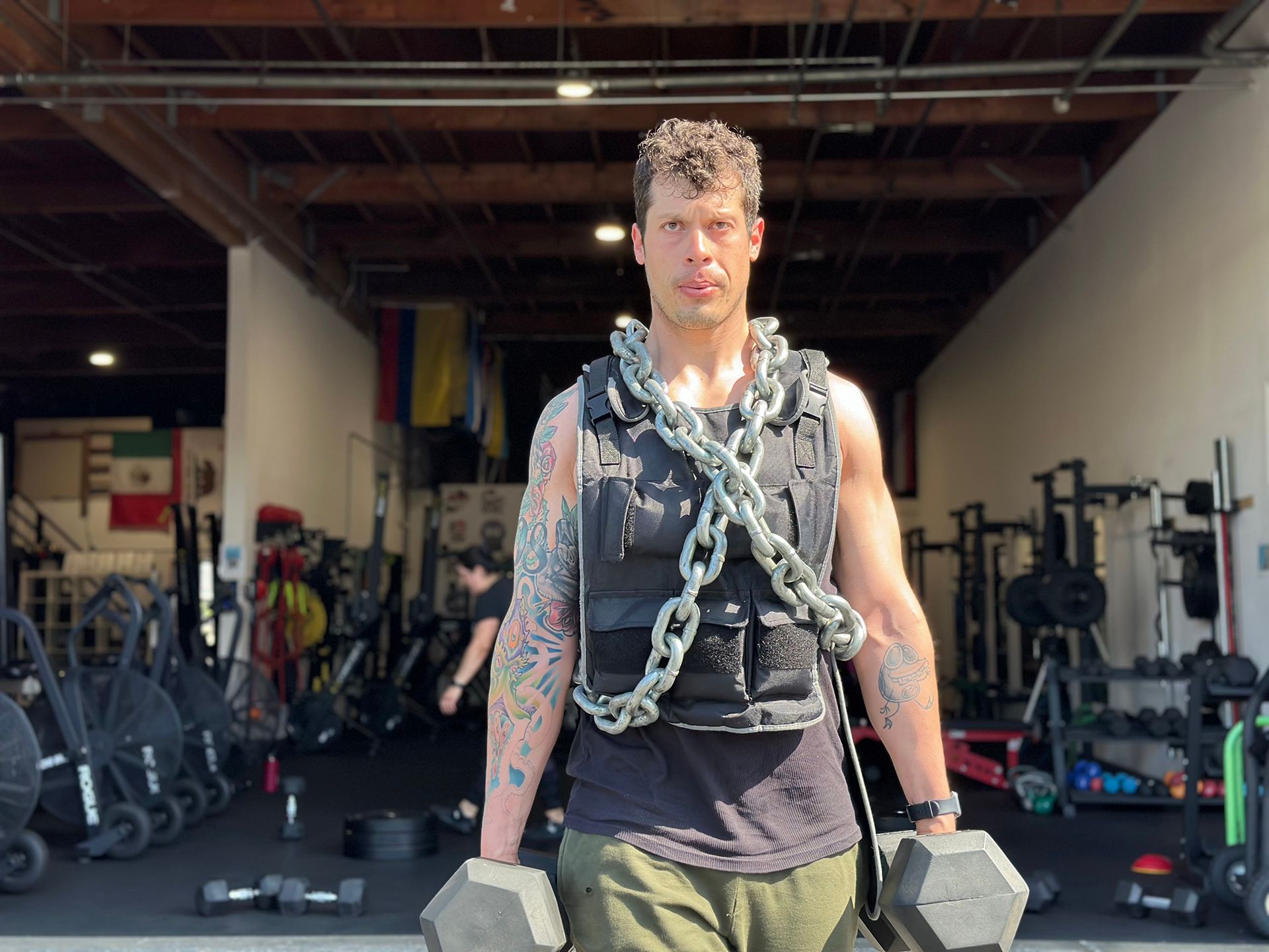 Man in a gym with weighted vest and dumbbells. He has tattoos and is looking forward.