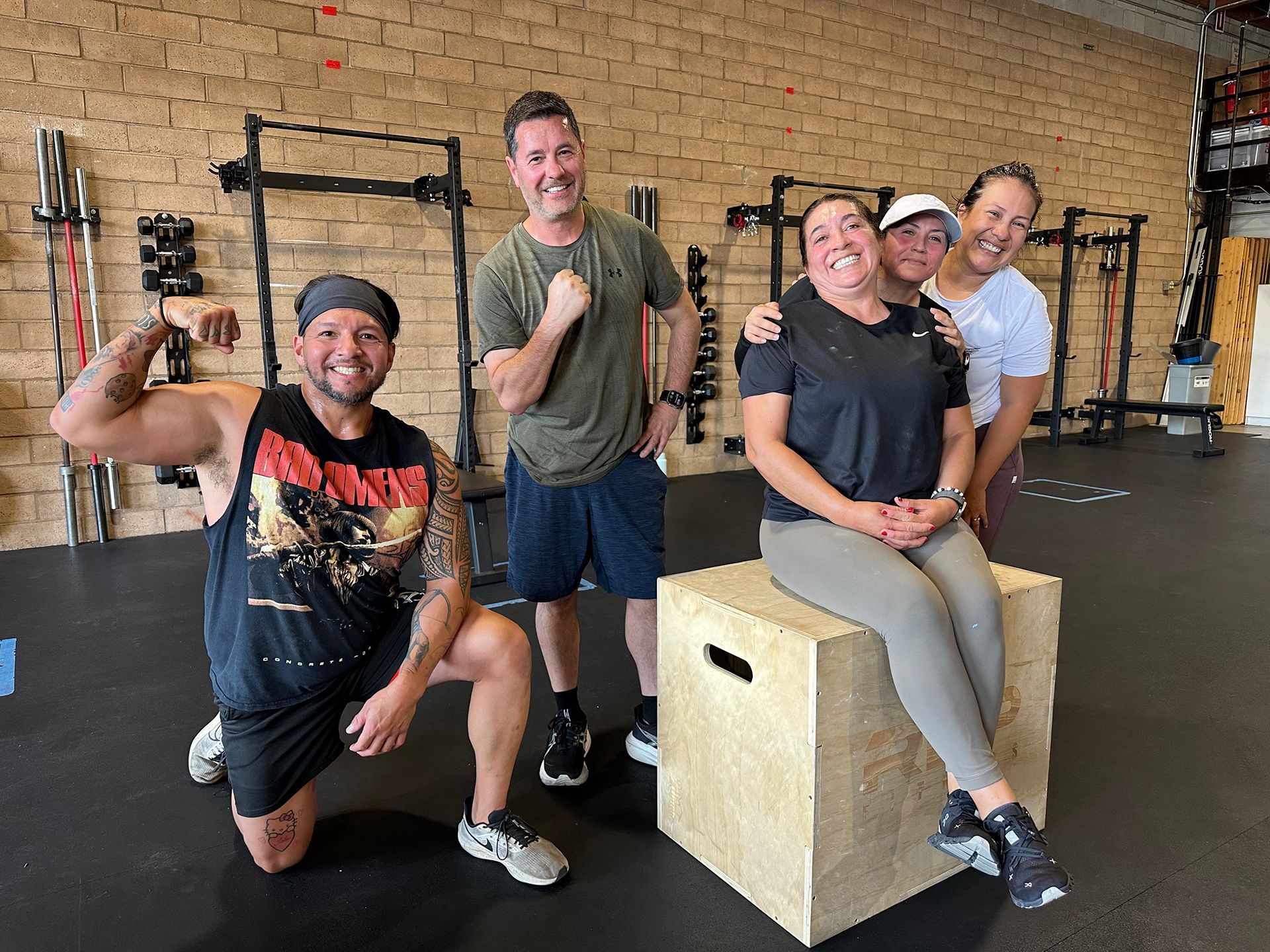 Group of five smiling people in a gym posing; one man flexes arm, one woman sits on wooden box.
