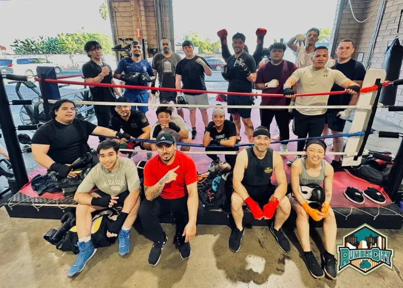 Group of boxers in a boxing ring, some raising fists. Bright setting, mixed ethnicity, gloves on, smiling.