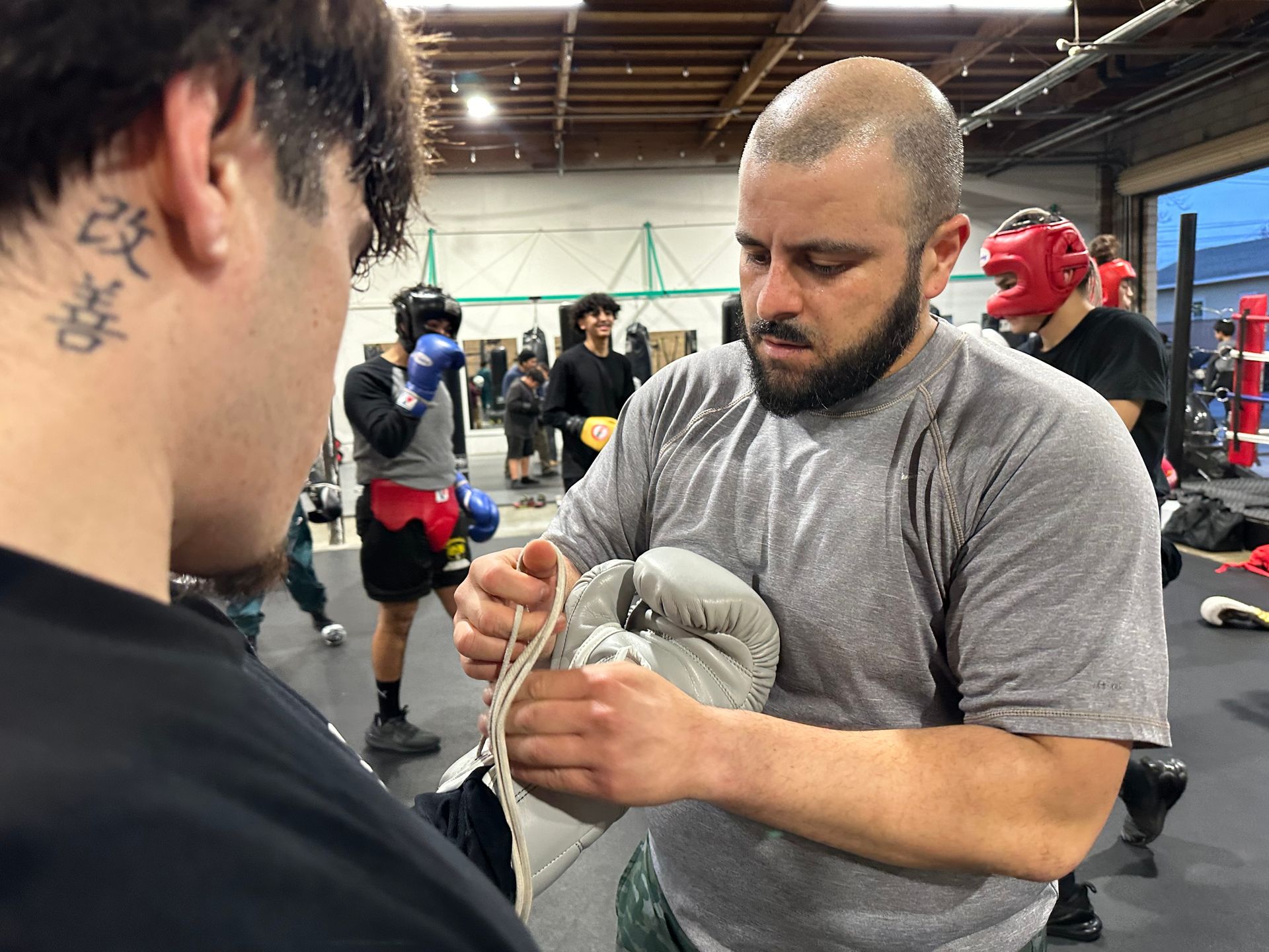 A man helping another put on a boxing glove inside a gym. Others train in the background.