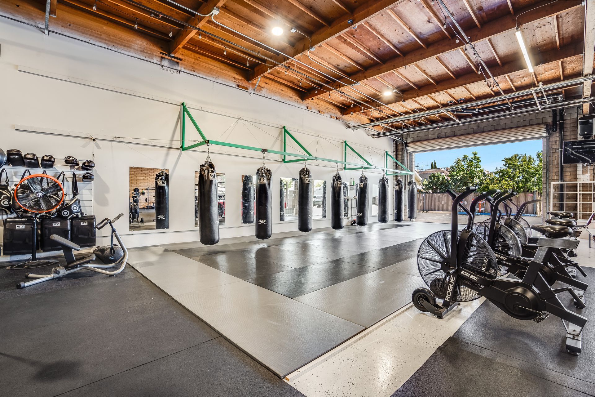 Gym interior with heavy bags, cardio equipment, and open garage door to outside.