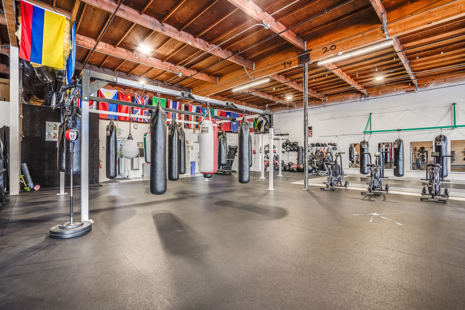 Boxing gym with heavy bags, equipment, and flags. Dark floor and wooden ceiling.