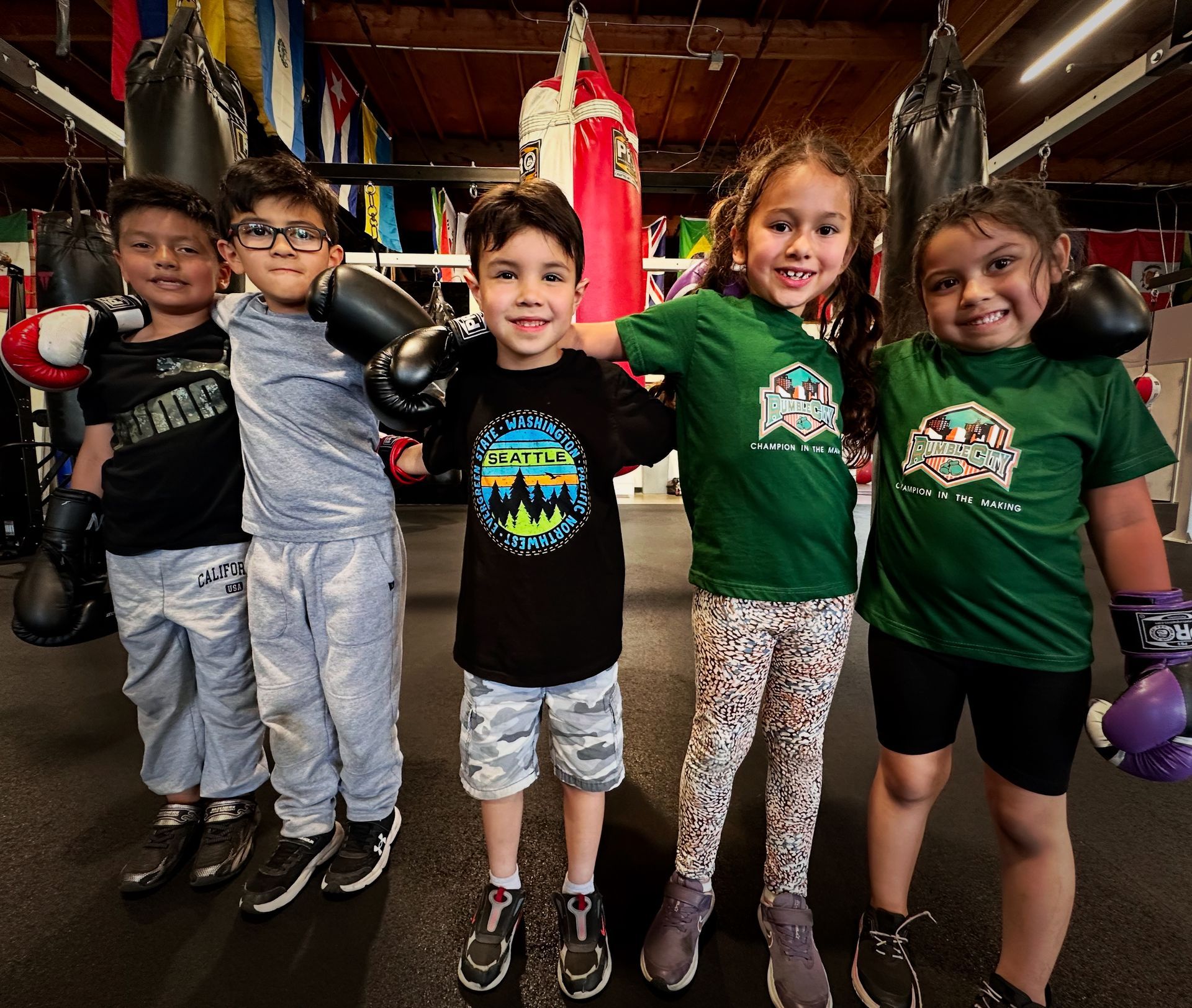 Five smiling children in a boxing gym, wearing gloves and boxing attire, posing together.