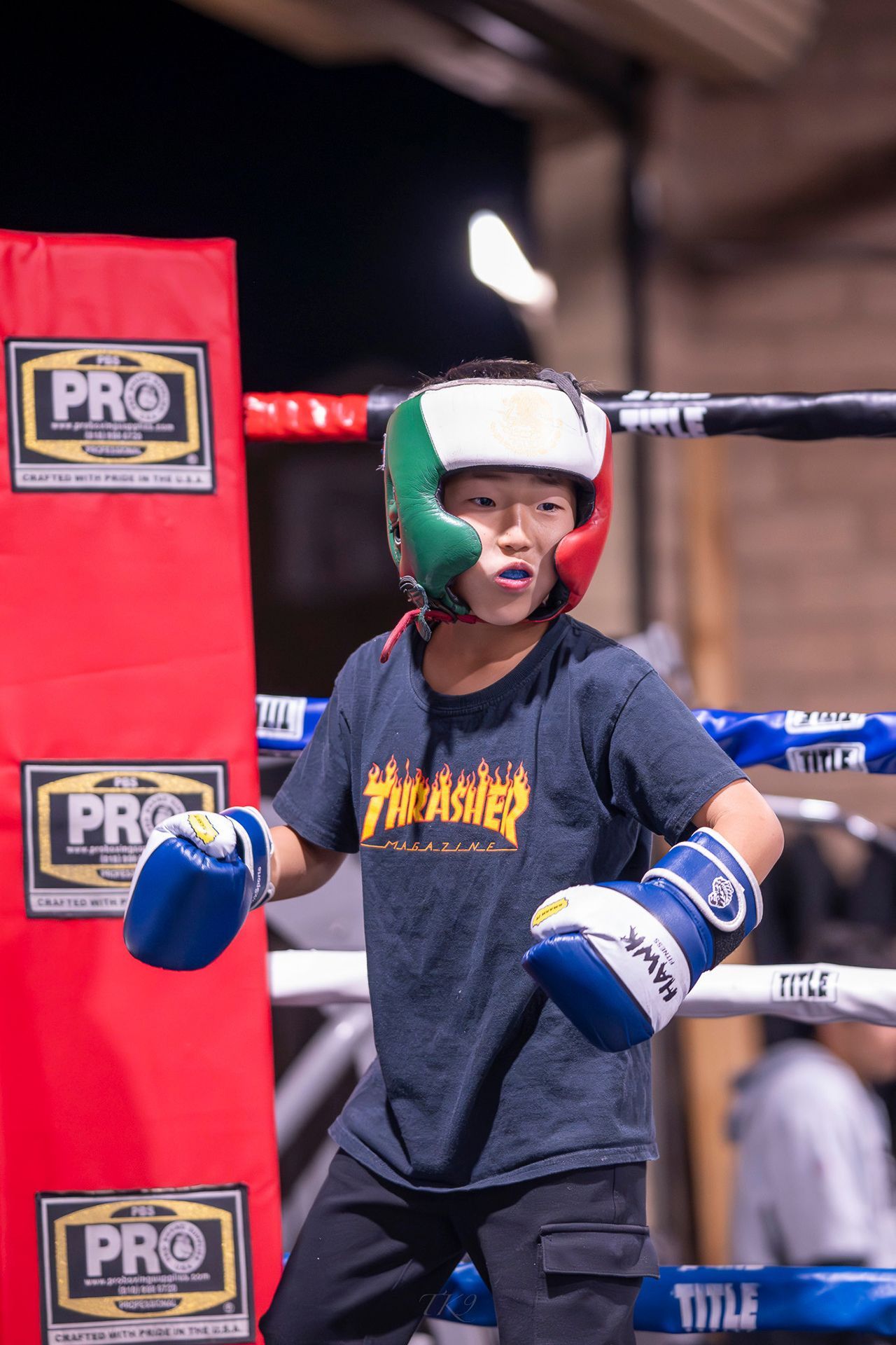 Young boxer in a ring, wearing a helmet, gloves, and a Thrasher shirt, looking focused.