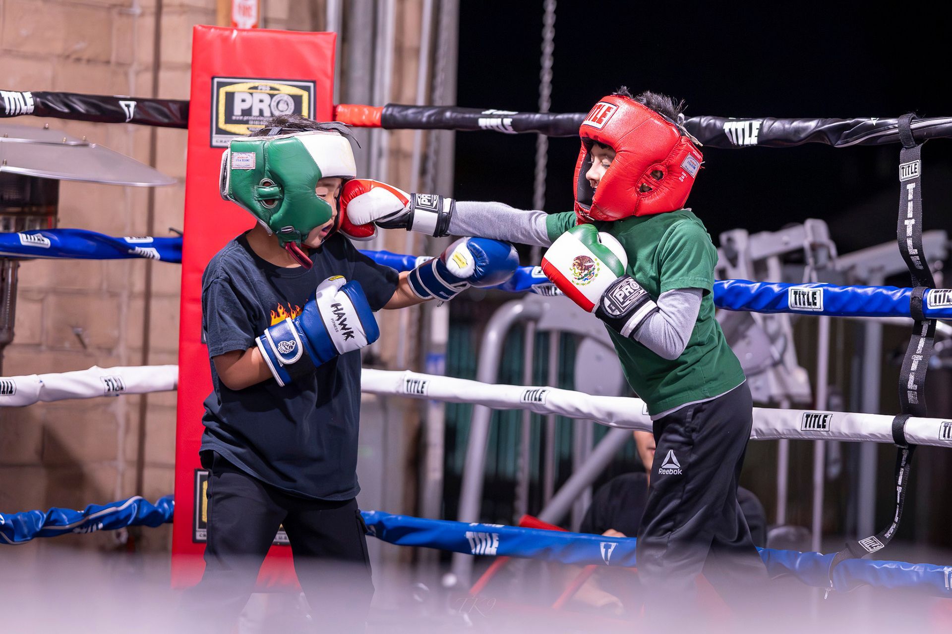 Two children boxing in a ring, wearing helmets and gloves; green and red colors, one throwing a punch.