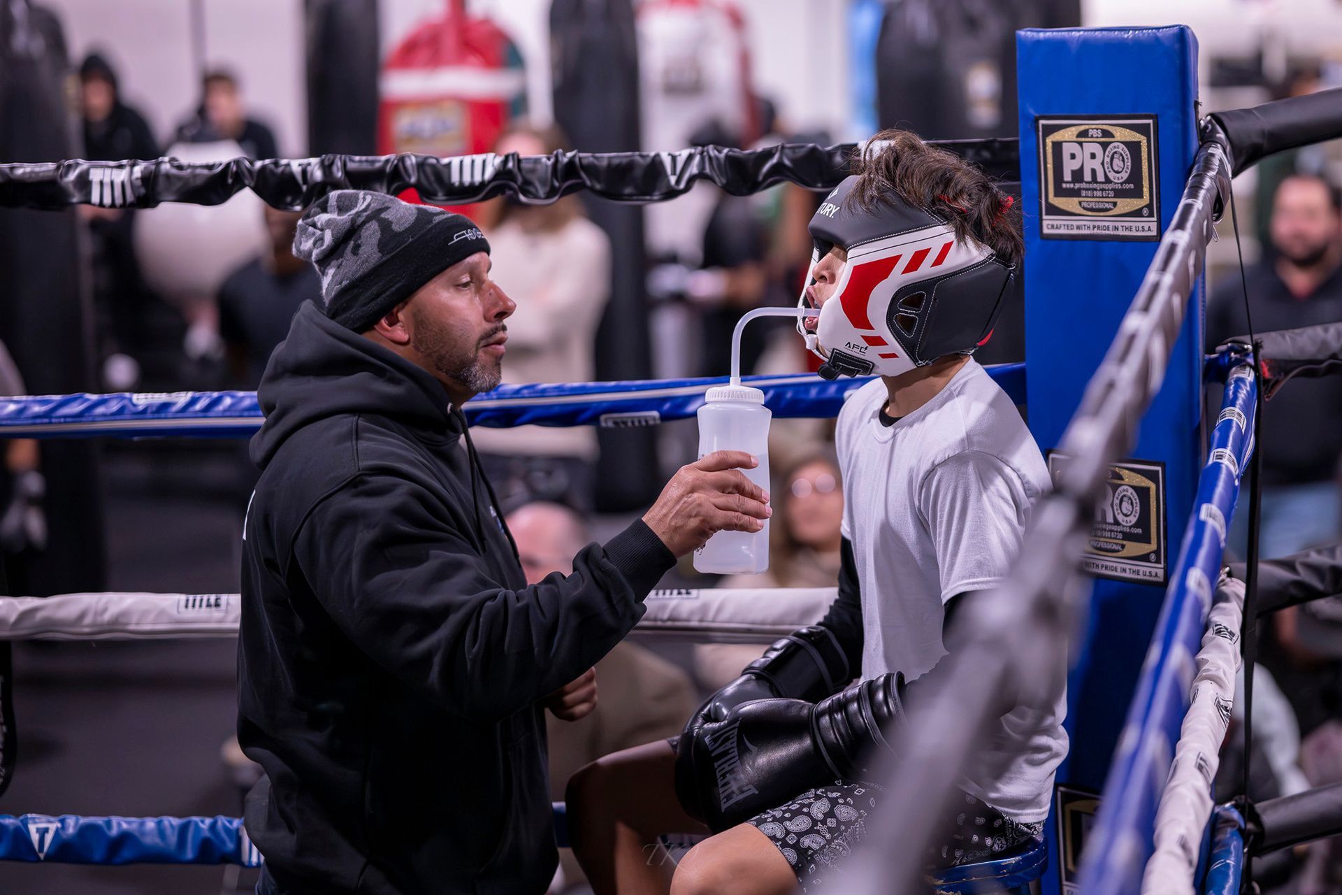 A boxing coach gives water to a young boxer in a ring, observing and guiding.