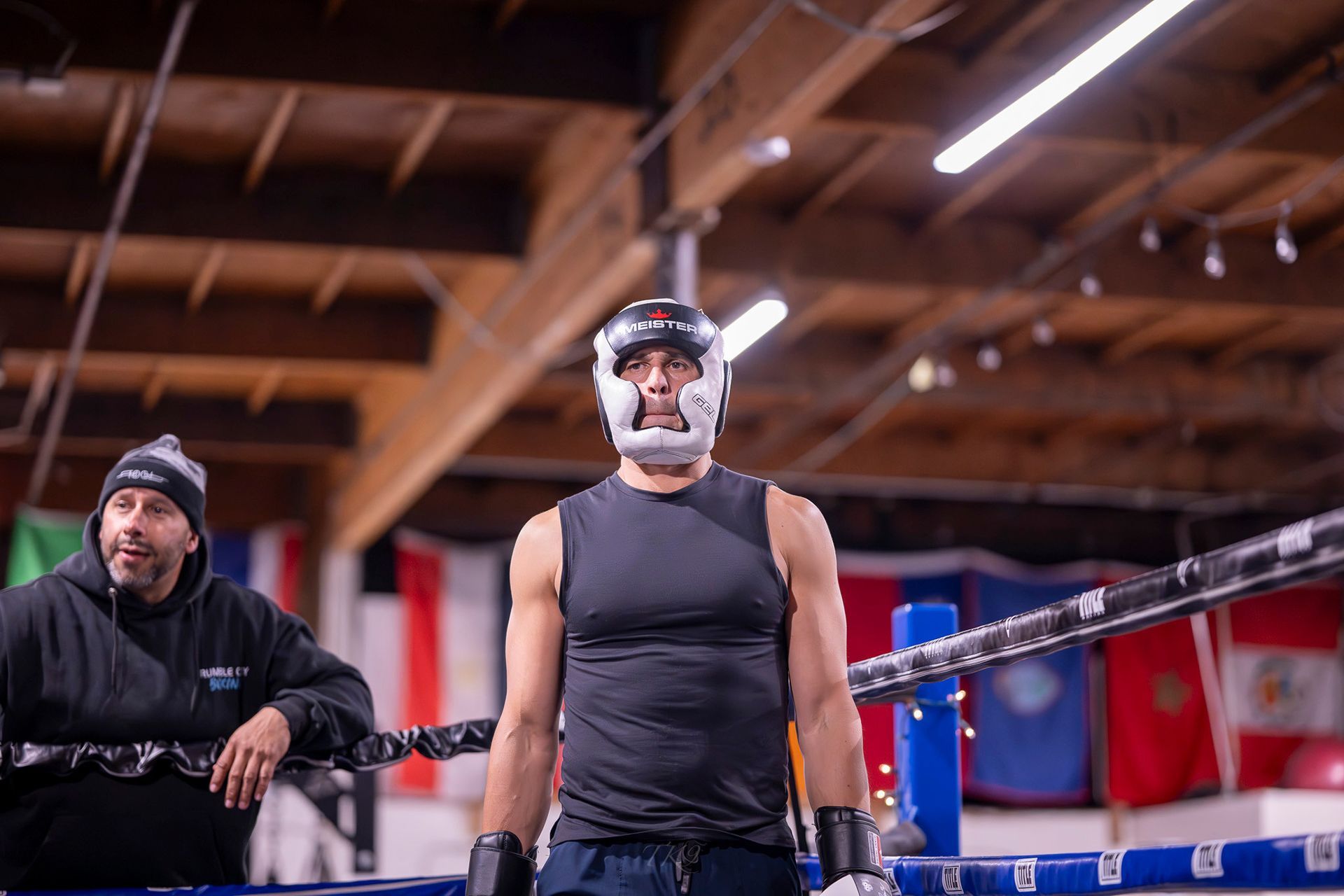 Two boxers sparring in a boxing gym, observed by others. Red brick walls, black floor.