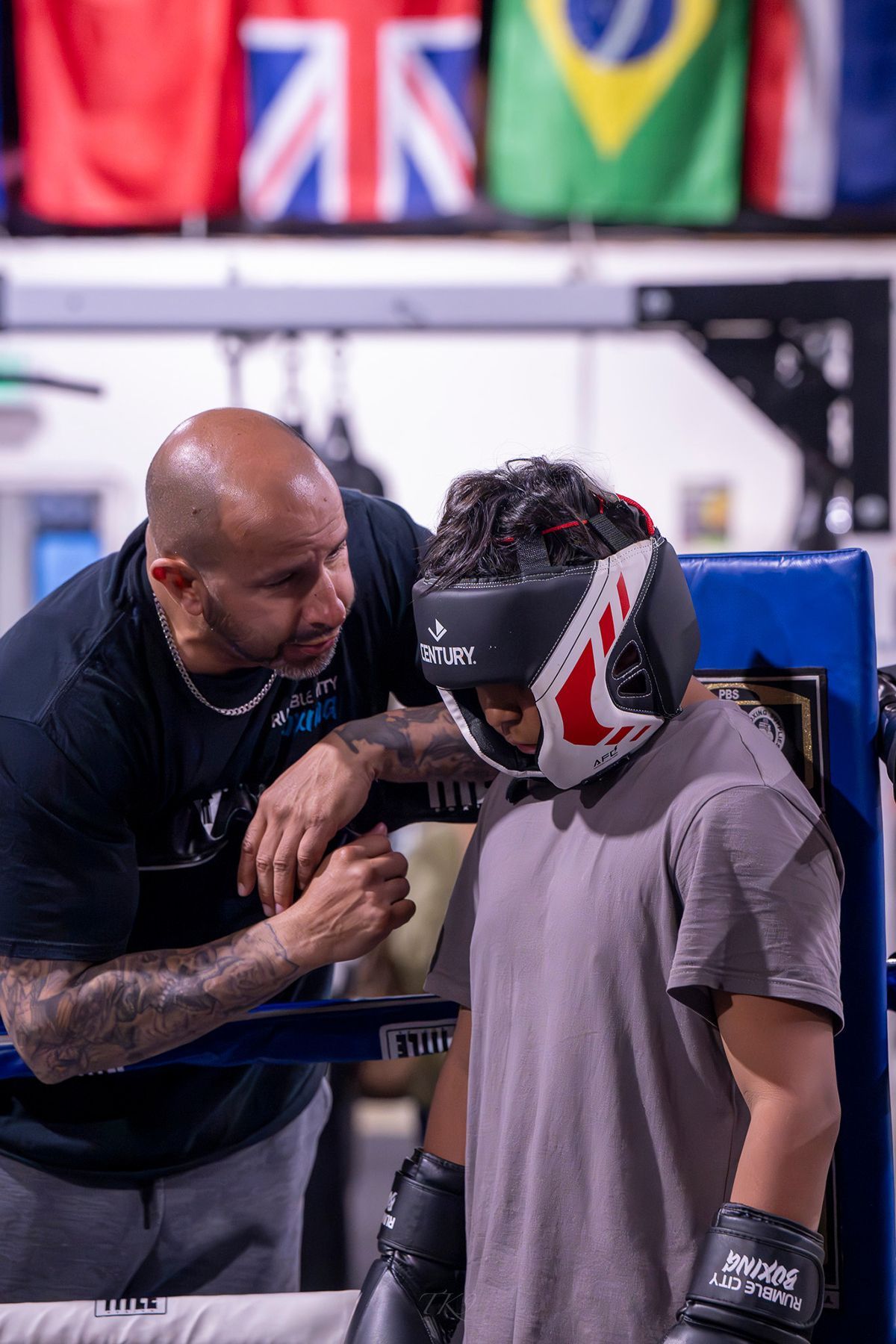 Boxer in headgear with coach in boxing ring. Coach has arm around boxer. Flags in the background.