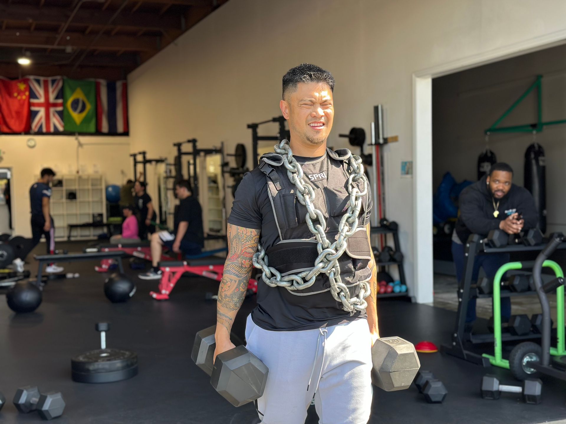 Man in gym with chains, weights, and arm tattoos, looking strained. Other people and equipment in the background.