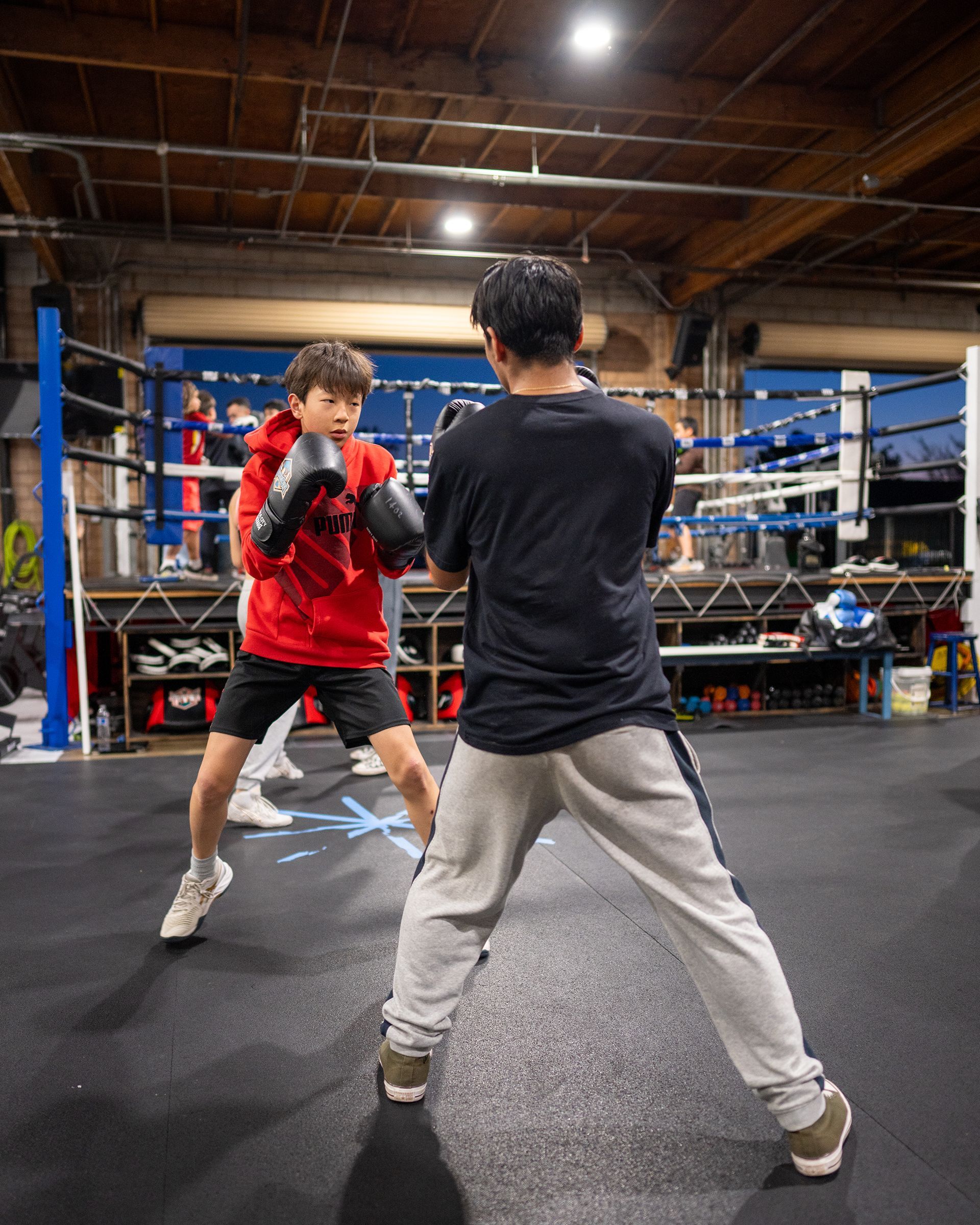 A young boxer in a red hoodie practices with a trainer in a boxing ring.