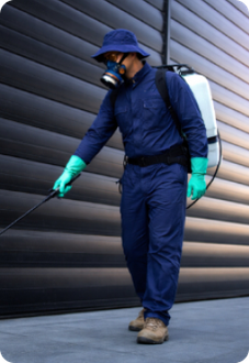 A professional in blue protective gear and a respirator walks while spraying a chemical solution along a dark wall.
