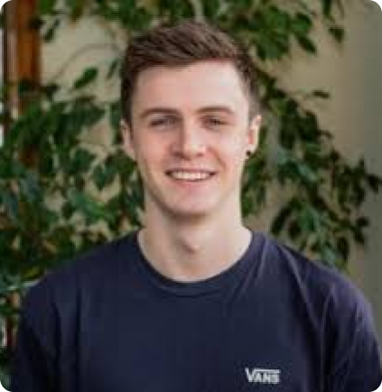 A smiling person wearing a dark navy Vans t-shirt, positioned in front of a blurred background of green leaves.