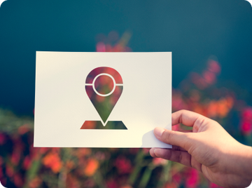 A hand holds up a white paper with a cut-out location pin icon against a soft, colorful, out-of-focus background.
