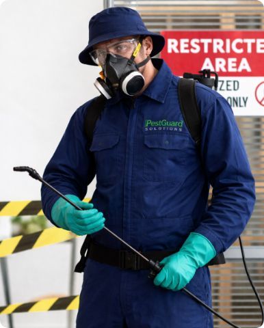 A pest control worker in blue uniform, bucket hat, and respirator holds a spray wand in front of a restricted area sign.