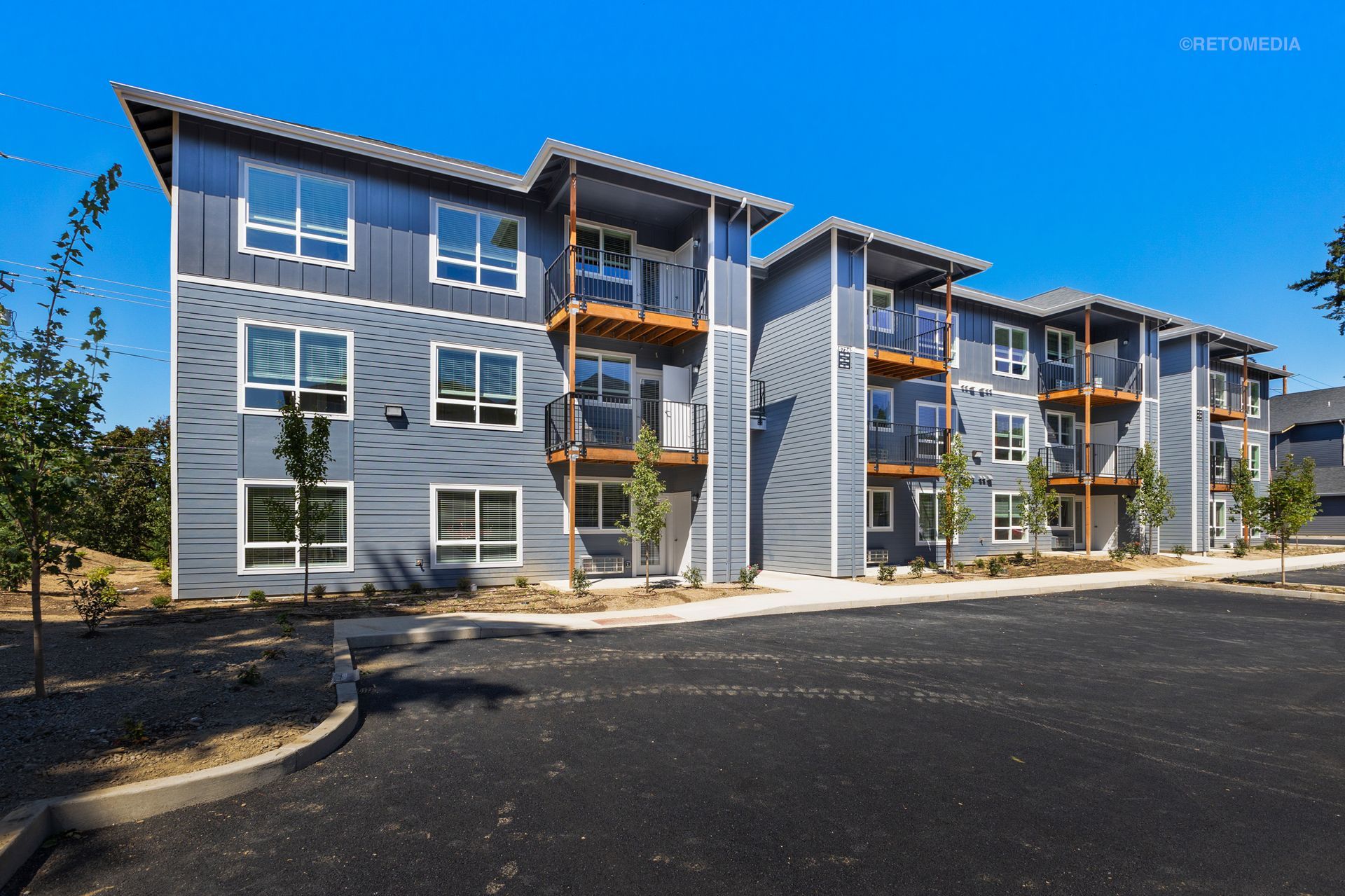 Modern gray apartment buildings with balconies and a paved parking area under a clear blue sky