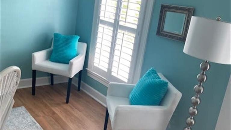 A living room with blue walls , white chairs , a lamp and a window.