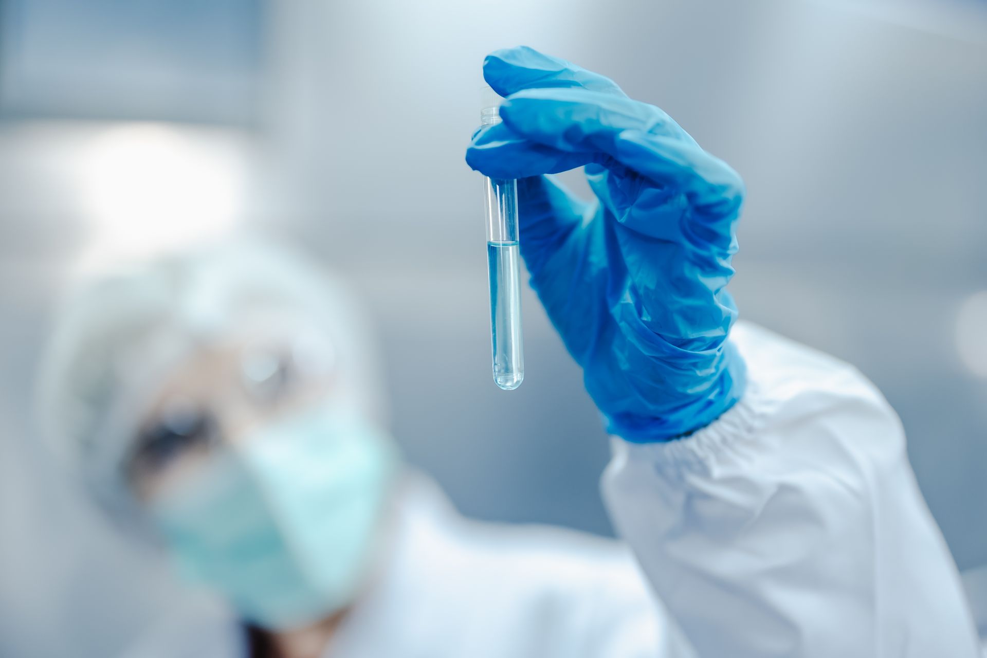 Scientist in lab coat and mask holding a test tube with blue liquid, wearing blue gloves.