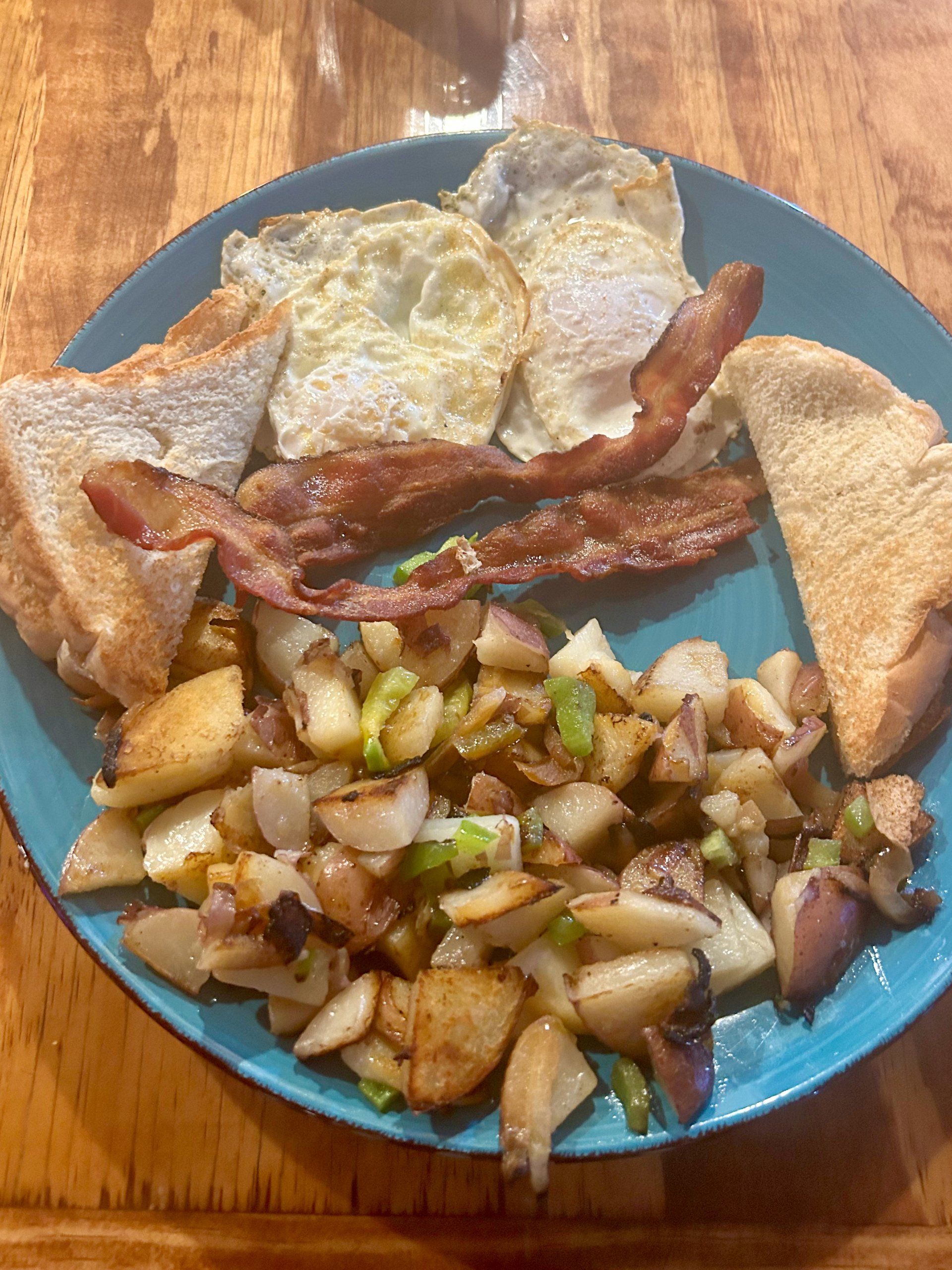 A blue plate topped with eggs , bacon , potatoes and toast on a wooden table.