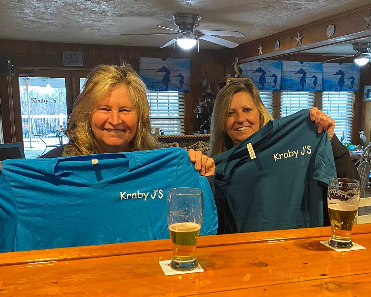Two women are sitting at a bar holding t-shirts that say kimberly j.s.