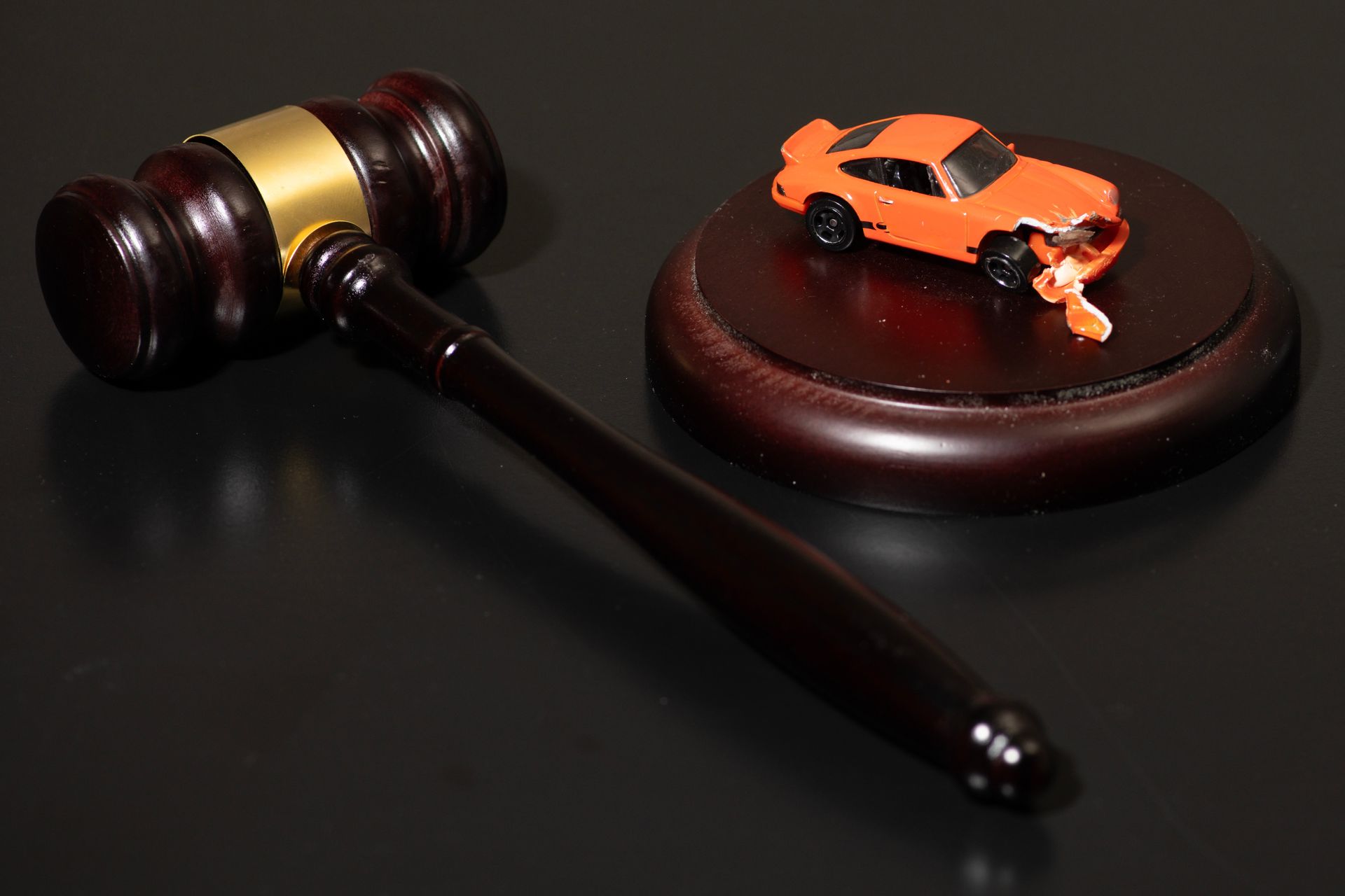Toy orange car on a round stand beside a judge’s gavel on a dark surface Toy orange car on a round stand beside a judge’s gavel on a dark surface