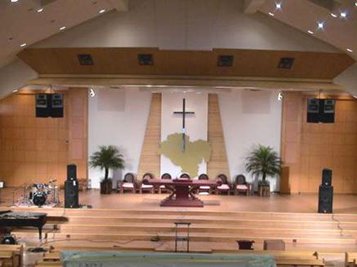 Church stage setup with cross, chairs, speakers, and palm plants on each side.