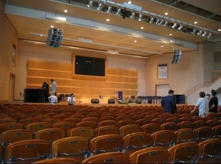 Large auditorium with ceiling-mounted speakers and rows of yellow chairs.