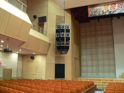 Line array speaker system hanging in a wooden auditorium above orange seats.