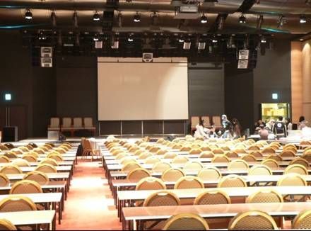 Large hall with rows of tables and chairs facing a stage with projection screen.