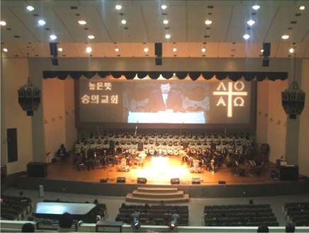 Church hall with choir on stage, projected sermon, and ceiling-mounted speakers.
