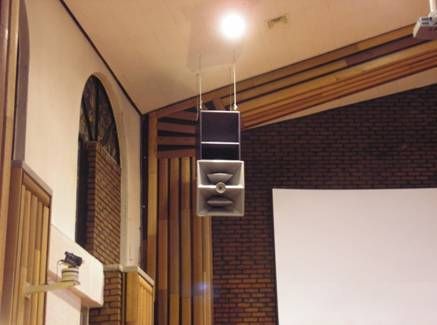 Ceiling-mounted speaker in a brick-walled church with wood acoustic paneling.