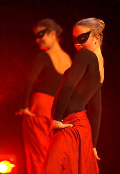 Masked dancers in red skirts and black tops performing under warm stage lighting.