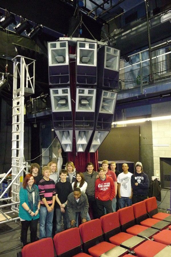 Students pose beneath suspended Funktion-One speakers in a theater space.