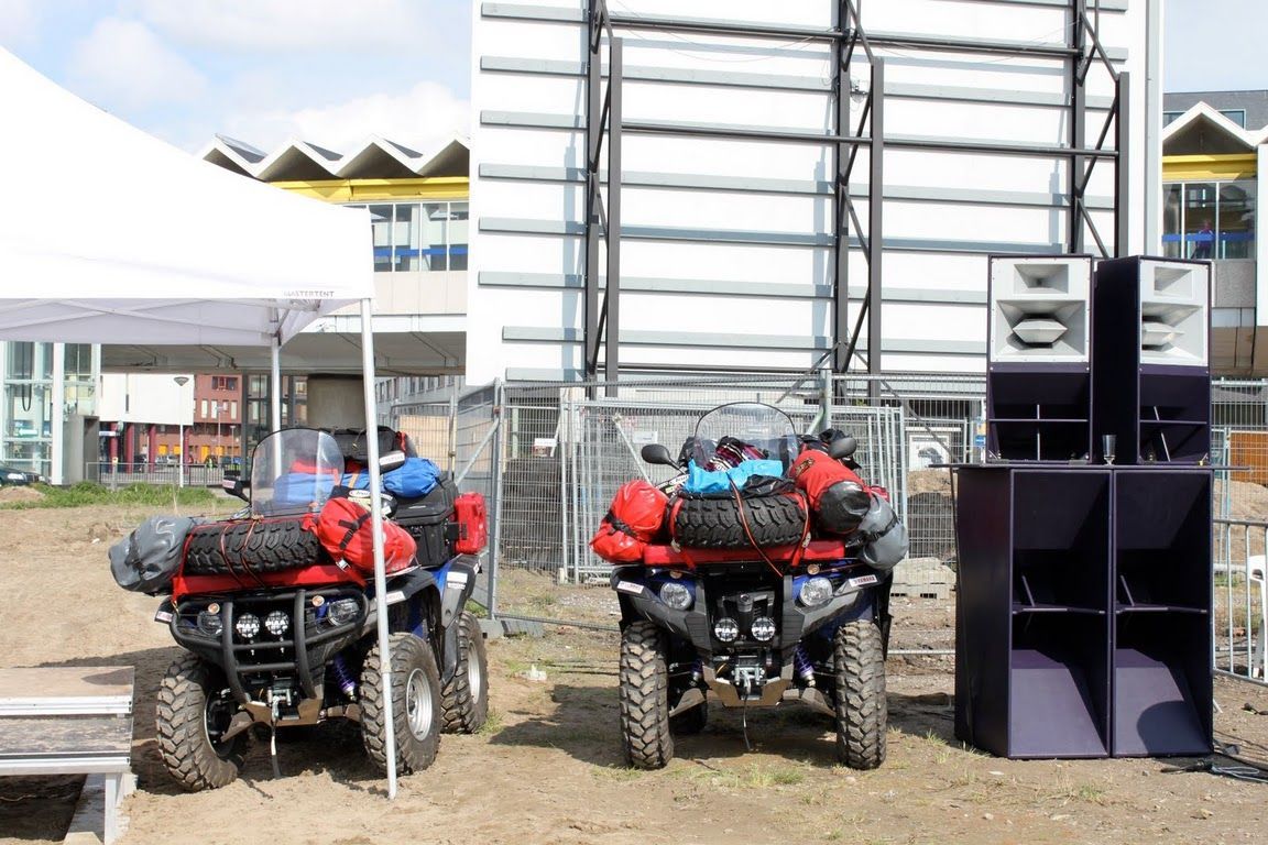 Two gear-loaded ATVs parked beside Funktion-One speaker stacks near city buildings.