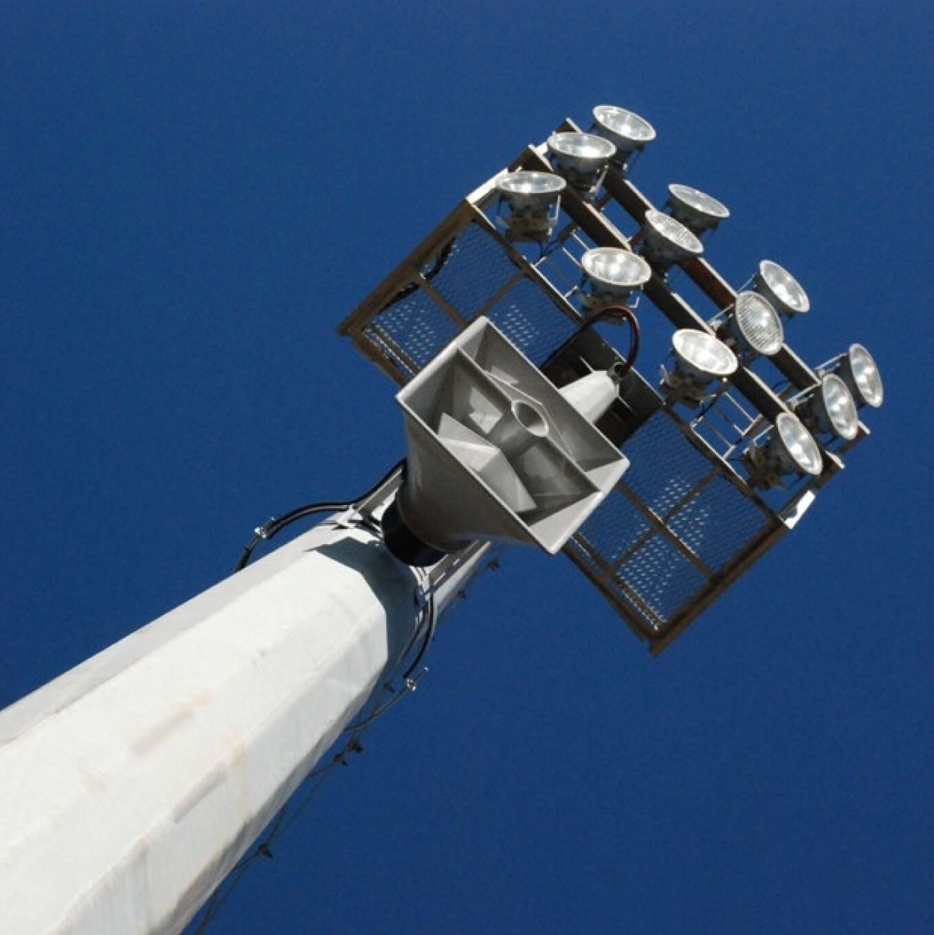 Looking up at a tall metal floodlight tower with multiple lights and a central speaker.