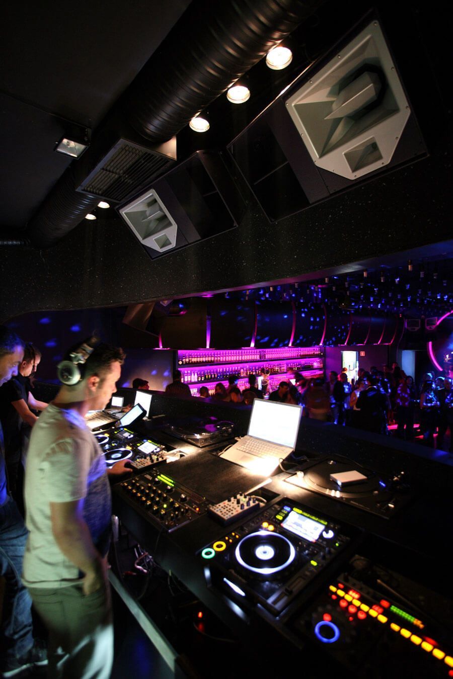 Club scene with a DJ performing live, an illuminated bar backdrop, and an energetic crowd on the dance floor.