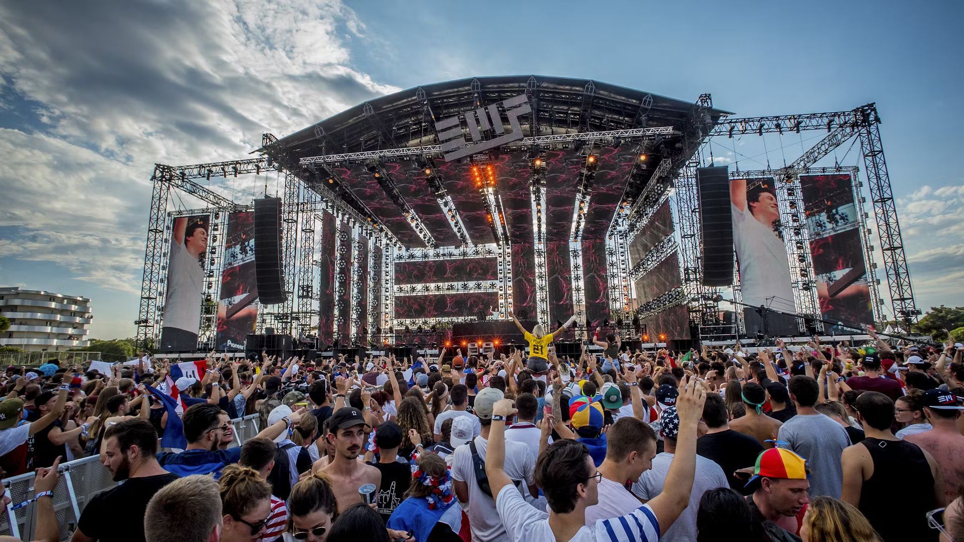 Massive crowd at outdoor EDM festival with lights, big screens, and large stage.