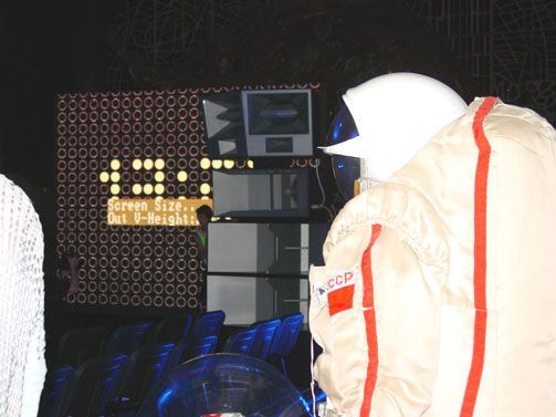Astronaut suit beside Funktion-One speakers and digital screen in dark venue.