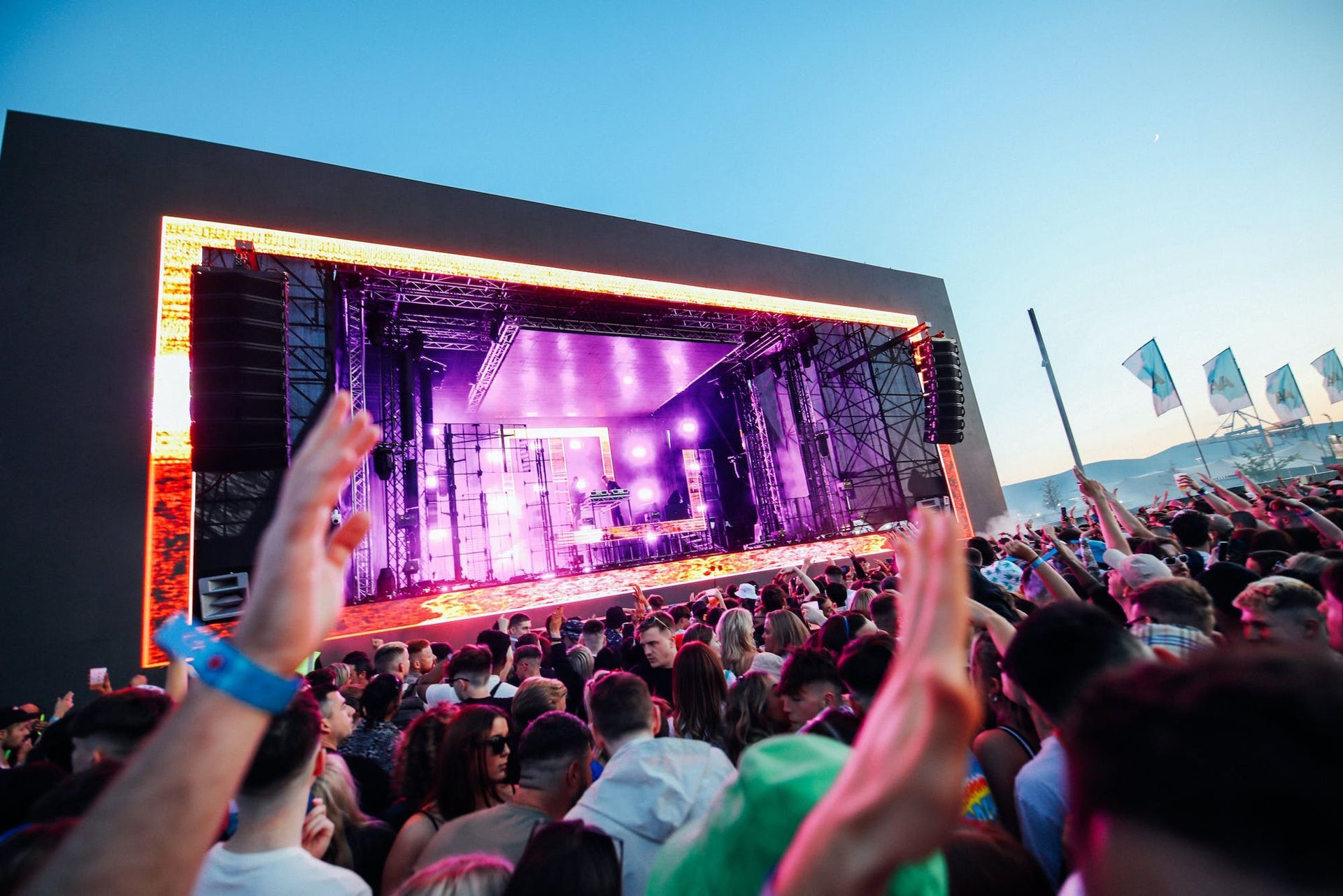 Crowd enjoying a live outdoor concert with bright lights and a DJ on stage during sunset.