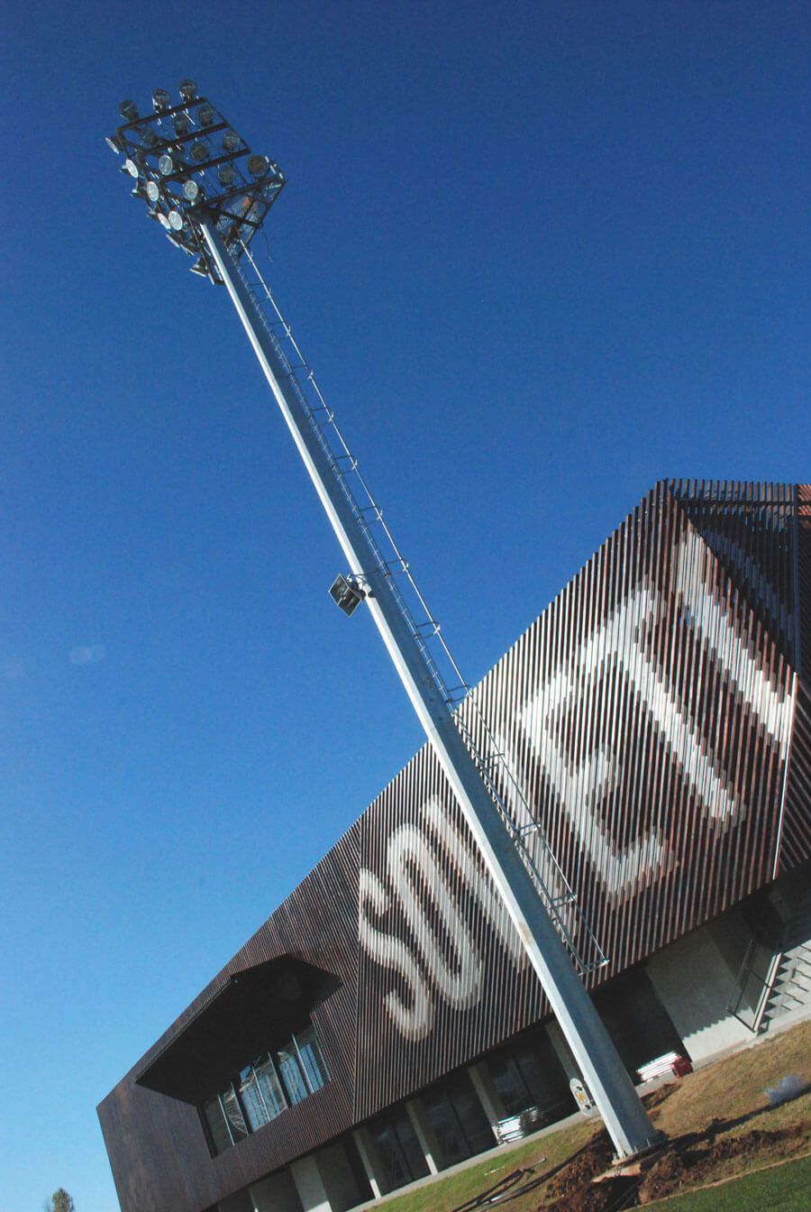 A tall metal light pole stands beside a modern building with vertical slats casting shadows. 