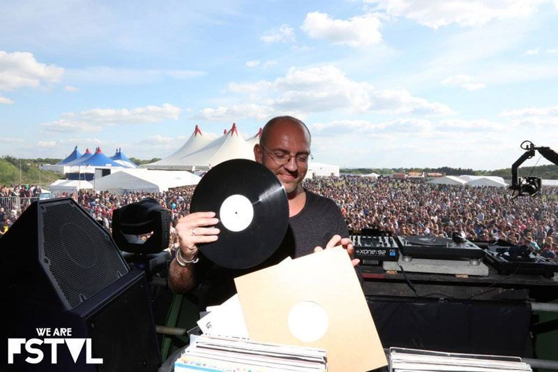 DJ holding vinyl at outdoor We Are FSTVL with tents and crowd in background.